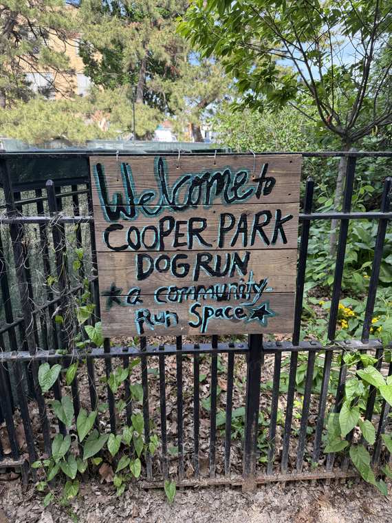 A hand-painted wooden sign zip-tied to the fence reading Welcome to Cooper Park Dog Run, a community run space