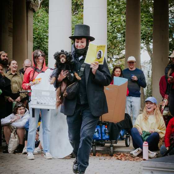 A parade entrant dressed as Abraham Lincoln holding a small dog in a matching costume, watched by a laughing crowd at the McGolrick pavilion